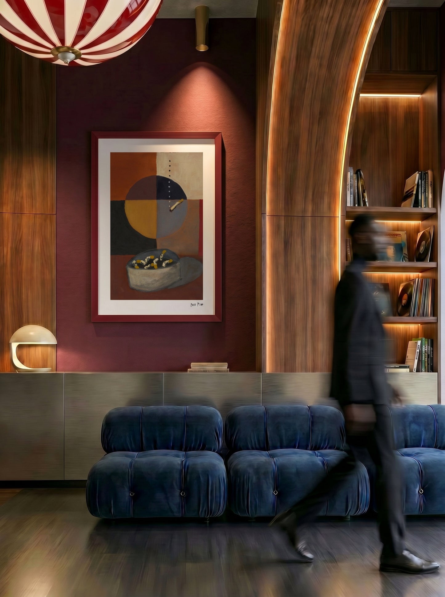 A framed abstract art print with geometric shapes hangs on a dark red wall in a modern interior. A blurred person walks in front of the artwork, and the scene includes blue velvet seating, wood paneling, built-in shelves, and warm lighting.