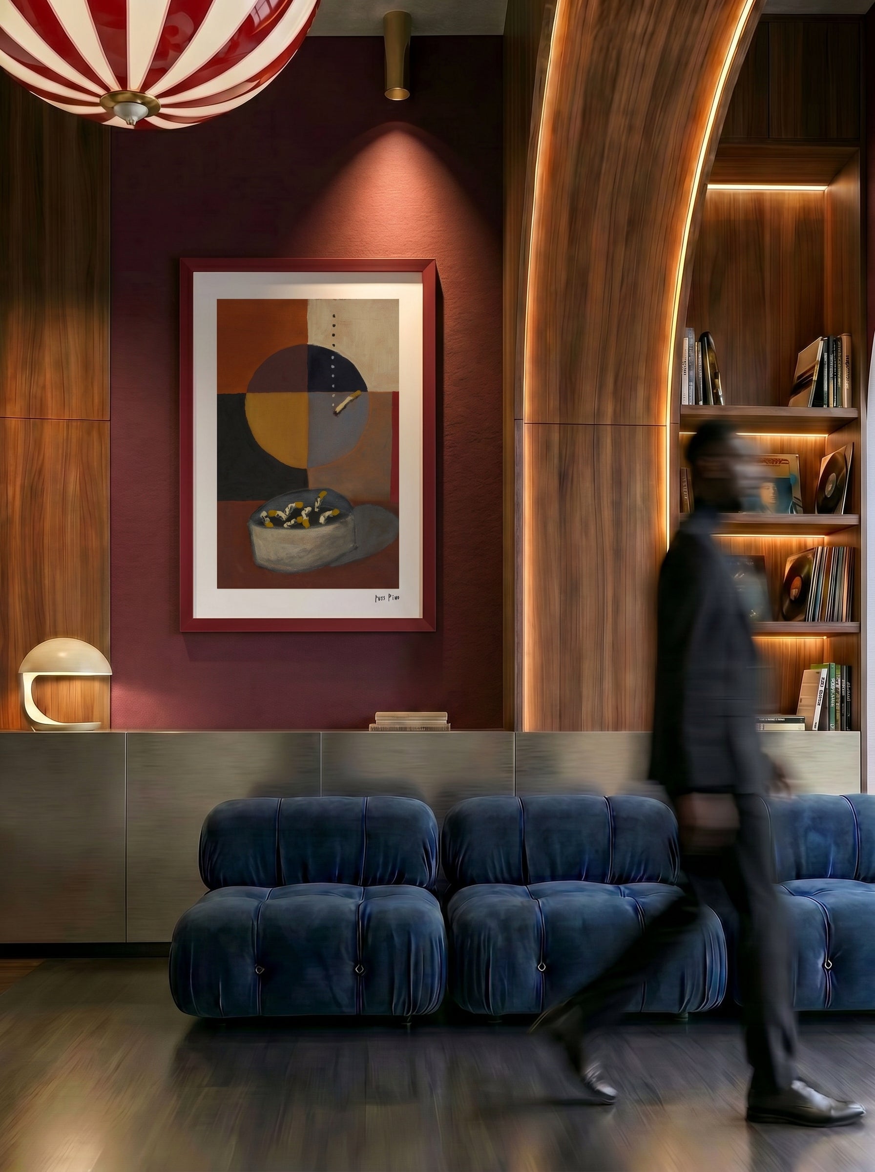 A framed abstract art print with geometric shapes hangs on a dark red wall in a modern interior. A blurred person walks in front of the artwork, and the scene includes blue velvet seating, wood paneling, built-in shelves, and warm lighting.